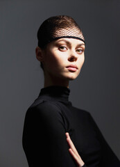 Thoughtful young woman with a fishnet headpiece, wearing a black long-sleeve top against a dark grey background. Concept of modern fashion and elegance.