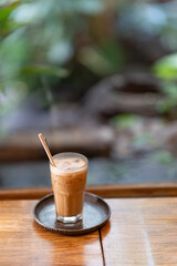 a glass of iced coffee on a rustic tray, placed on a wooden table with a softly blurred green background. The scene has a cozy, relaxed cafe ambiance.