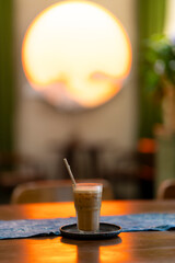 a glass of iced coffee on a tray with a straw, placed on a table with warm lighting. A large, glowing circular light in the background creates a cozy, sunset-like ambiance.