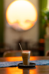 a glass of iced coffee on a tray with a straw, placed on a table with warm lighting. A large, glowing circular light in the background creates a cozy, sunset-like ambiance.