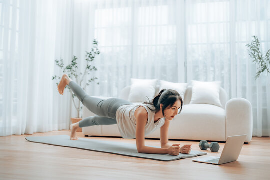 Young Asian woman in sportswear doing plank on training mat while watching online workout tutorials on laptop in the living room. Sport and recreation concept
