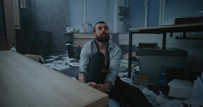 Dolly shot of stressed businessman sitting on the floor after fit of rage over failed business deal or receiving bad news. Depressed office employee in destroyed office room. Job burnout or firing.
