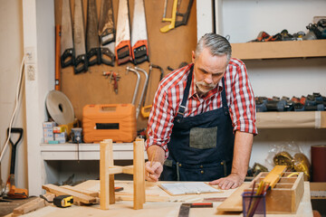 Man in plaid shirt and overalls working on a wooden project in his workshop, drawing on clipboard...