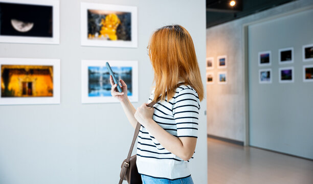 Visitor woman standing takes picture art gallery collection in front framed paintings pictures with mobile phone, Asian female watching at photo frame with smartphone at artwork gallery show