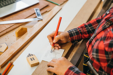 Person in a red plaid shirt sketching table plans on paper with a red pencil, surrounded by wooden planks and miniature houses. Perfect for architecture, construction, and DIY project concepts