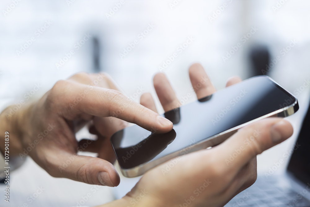 Poster Focused image of a man navigating his high-tech smartphone, laptop open next to him amidst the blur of a busy office setting