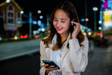 A young woman talking on her mobile phone with earphones while walking on a city street at night....