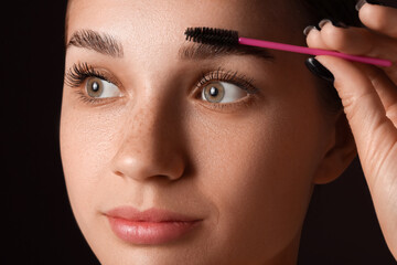 Young woman correcting shape of her eyebrows on dark background, closeup