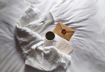 A white blanket, a cup of coffee, an envelope, and a book lie on a white bed.