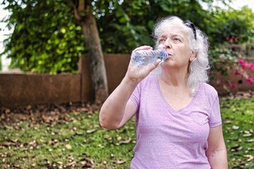 Gray-haired elderly lady drinks water from a bottle in the park.