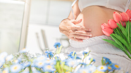 Pregnant woman sitting finger touching stomach, belly and Holding a flower in the other hand, catching the sunlight among the flowers. Concept of love from mother
