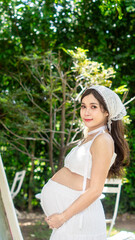 Beautiful Asian pregnant woman in white dress wearing bohemian style headdress Hug her belly at the outside park, playground