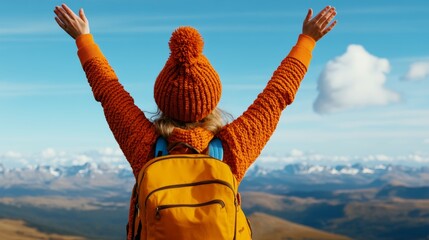 Woman wearing a bright orange jacket and a red hat is standing on a mountain top, looking up at the sky. She is holding her arms up in the air, as if she is celebrating something