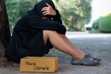 Asian male teenager wearing black headscarves sits on floor near tree in public park as he was...