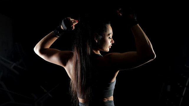 The silhouette of a female bodybuilder on a black background, she stands with her back to the camera and shows muscular arms and back. Portrait of a sporty woman in a dark gym.