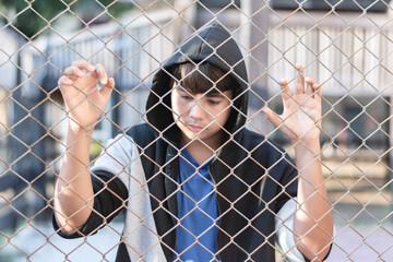 A young boy wearing a gray hoody stands with his hands on a metal fence in a local detention...