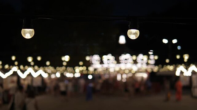 The blurred carousel rotates and glows with lights in the evening park. A blurry view of a carousel in an amusement park in the evening, people walking along a glowing alley.