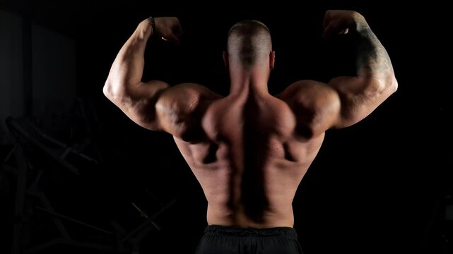 A strong, semi-naked man stands with his back to the camera and shows off his muscles in a dark room. The bodybuilder poses with his back to the camera, the shadow on his body shows every muscle.