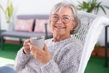 Portrait of senior attractive woman with cheerful smile relaxing at the armchair in the balcony at home looking at camera holding a coffee cup