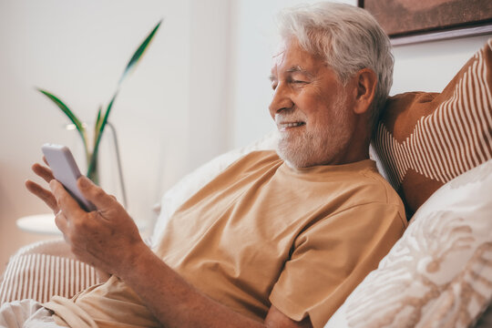 Portrait of handsome smiling senior bearded man sitting relaxed in bed using phone enjoying tech and social. Old people and modern activity