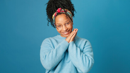 Smiling young woman with glasses dressed in blue sweater, pressing her palms to her face, looks into camera, tenderness, on blue background in studio
