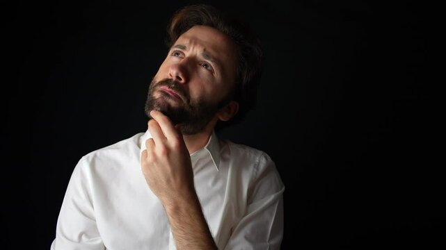 A young Caucasian man in a white shirt shows visible uncertainty and doubt, his facial expression shifting as he touches his chin. Black background. 