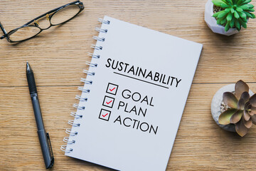 Fototapeta premium Sustainability written in notebook with pen, glasses and two small plant on wooden table. business sustainable concept.