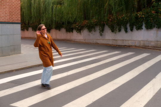 Person walking on a pedestrian crosswalk in urban environment near a building and greenery. Street photography with motion blur. 50 year old woman (fifty years old)
