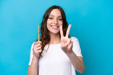 Young caucasian woman brushing teeth isolated on blue background smiling and showing victory sign