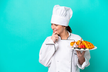 Young caucasian chef woman holding waffles isolated on blue background thinking an idea and looking side