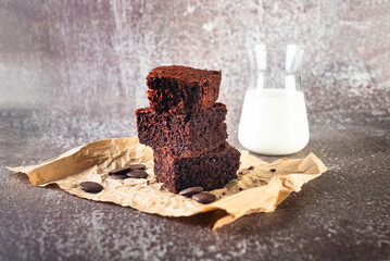 A stack of chocolate brownies on bakery paper and glass of milk on dark background, homemade cakes...