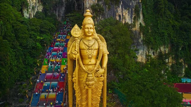 A towering golden statue of a Hindu deity stands majestically at Batu Caves in Malaysia, while numerous visitors ascend the vibrant, multicolored steps leading to the caves.