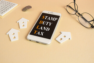 Concept words SDLT-Stamp Duty Land Tax on the mobile phone screen. Beautiful beige background. Glasses, miniature houses, coins. Buying housing and land. Property concept.