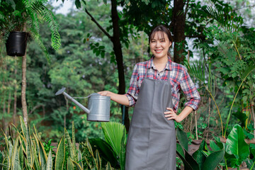 Asian woman watering plants in lush garden. Wearing apron and plaid shirt, surrounded by green foliage and potted plants. Smiling, enjoying weekend gardening outdoor activity, garden working in botany