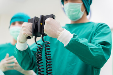 Close-up of Asian surgeon in green scrubs holding defibrillator paddles in operating room. Emphasis...