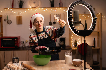 baker online courses. Smiling female blogger cooking gingerbread cookies and shooting video of herself using mobile phone on a tripod. Culinary training class