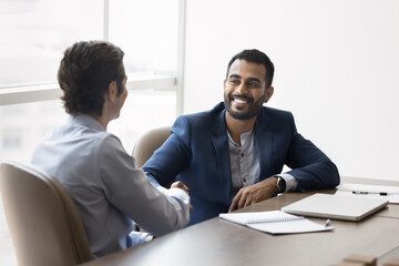 Happy young Indian business owner man shaking hands with male partner at meeting table, hiring new...