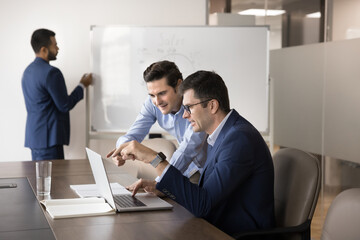 Two diverse business coworker men watching Internet project presentation, website content on laptop in boardroom, speaking, smiling, pointing at computer monitor, meeting at desktop for teamwork