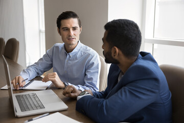Obraz premium Two serious multiethnic businessmen talking at laptop, using online modern technology for teamwork, communication, discussing Internet startup, cooperation strategy, sitting at meeting table