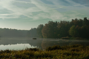 Jezioro położone w wysokim, sosnowym lesie. Jest jesienny poranek. Nad jeziorem unosi się mgła rozświetlana promieniami słońca. © boguslavus