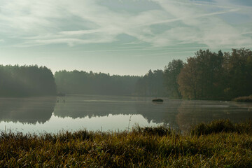 Jezioro położone w wysokim, sosnowym lesie. Jest jesienny poranek. Nad jeziorem unosi się mgła rozświetlana promieniami słońca. © boguslavus