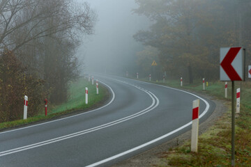 Asfaltowa droga prowadząca przez las, w mglisty, jesienny poranek. Nad jezdnią unosi się mgła ograniczająca mocno widoczność. © boguslavus