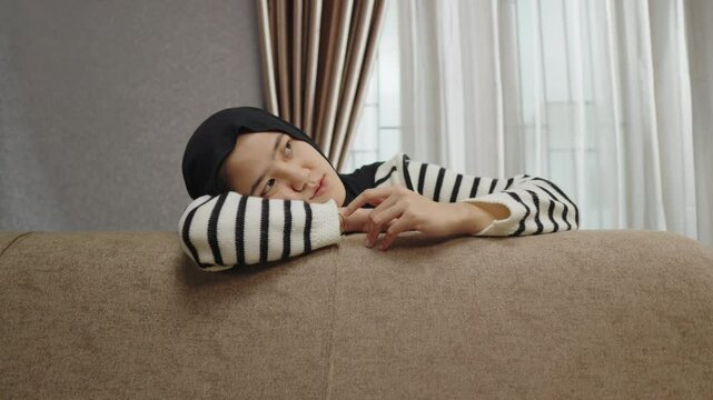 A portrait of a Muslim woman feeling stressed and anxious, resting her head on the sofa. A tired and confused expression due to overwhelming worry and unresolved mental burdens.