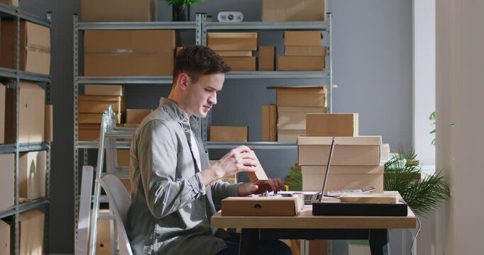 Warehouse worker carefully matches order numbers on cardboard boxes to online invoices using laptop. Side view of concentrated man working on laptop while sitting among shelves with cardboard boxes.
