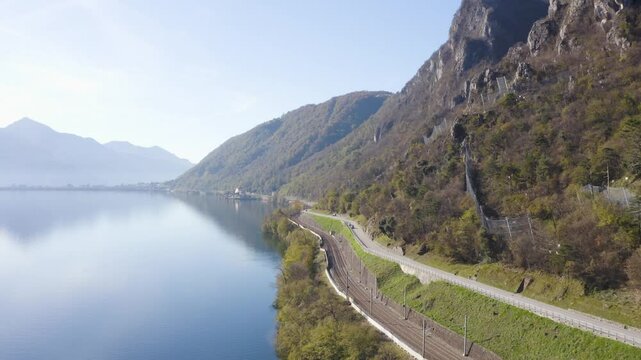 Drone flies over Lake Ceresio in Switzerland in the direction of Melide. On the right you can see the roadway and the railway.