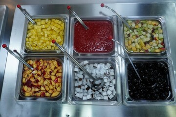 Toppings for yogurt, ice cream or other desserts. Containers with assorted fruits. Cubes of yellow fruits, pineapple in syrup or juice. Fruit salad. Thick red puree. Strawberry or other berry sauce