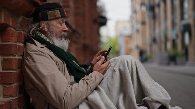 Side view of unhappy elderly homeless man sitting by brick wall using smartphone, possibly seeking help or connecting with family. Aged tramp male using phone to seek work and government assistance.