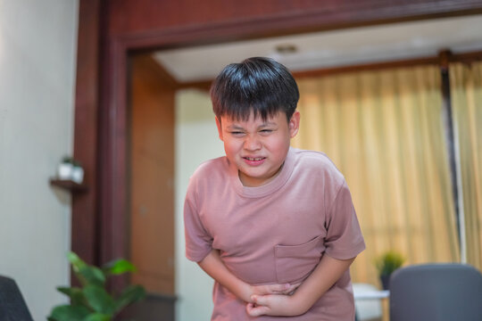 Portrait of sick unhappy little child asian boy touching belly stomach with suffering from stomach ache pain, gastritis, gastroenteritis, food poisoning, diarrhea, intestinal inflammation at home