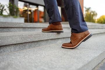 Low section of senior male entrepreneur in brown leather shoes moving up on staircase in modern city