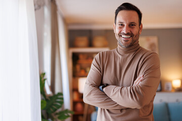 Portrait of positive handsome man with arms crossed smiling at camera and standing by window at home
